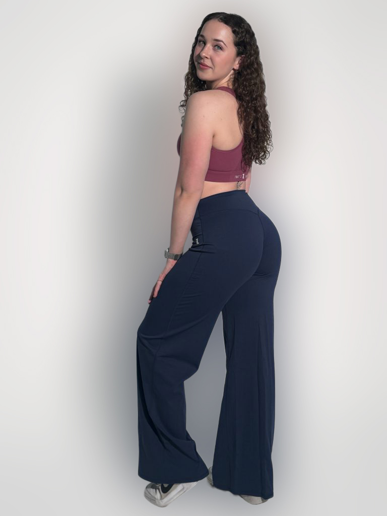 A woman with curly hair, wearing a mauve sports bra, navy blue flared pants, and white sneakers, stands sideways and looks back at the camera against a plain white background.