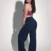 A woman with curly hair, wearing a mauve sports bra, navy blue flared pants, and white sneakers, stands sideways and looks back at the camera against a plain white background.