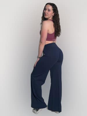 A woman with curly hair, wearing a mauve sports bra, navy blue flared pants, and white sneakers, stands sideways and looks back at the camera against a plain white background.