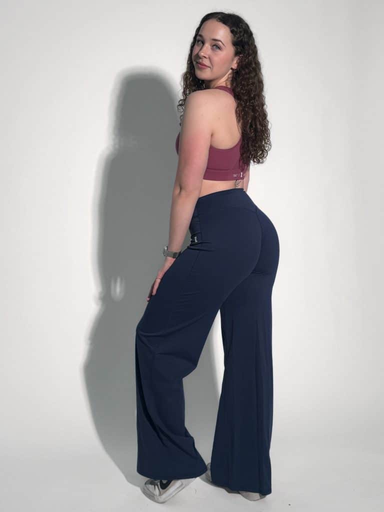 A woman with curly hair, wearing a mauve sports bra, navy blue flared pants, and white sneakers, stands sideways and looks back at the camera against a plain white background.