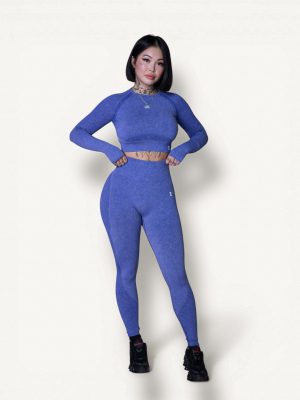 A woman with a bob haircut wearing a matching blue long-sleeve crop top and leggings, black sneakers, and a necklace stands confidently with hands on her waist against a plain white background.