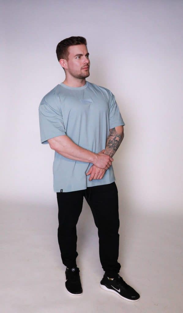 A man with short brown hair and a trimmed beard stands against a plain background wearing the Equity Tee, featuring a tapered fit and scoop bottom, along with black pants and sneakers. His arms are crossed as he looks to the side.