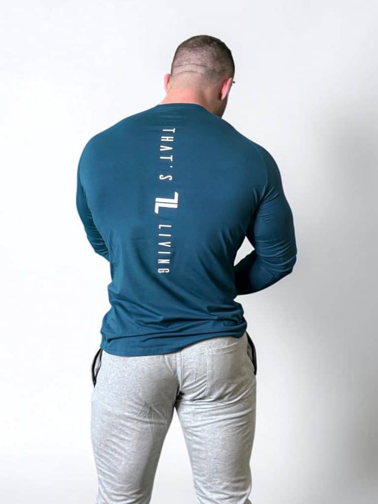 A muscular man stands facing away, wearing a fitted teal long-sleeve shirt with "THAT'S LIVING" and a logo printed vertically on the back, and light gray joggers, against a plain white background.