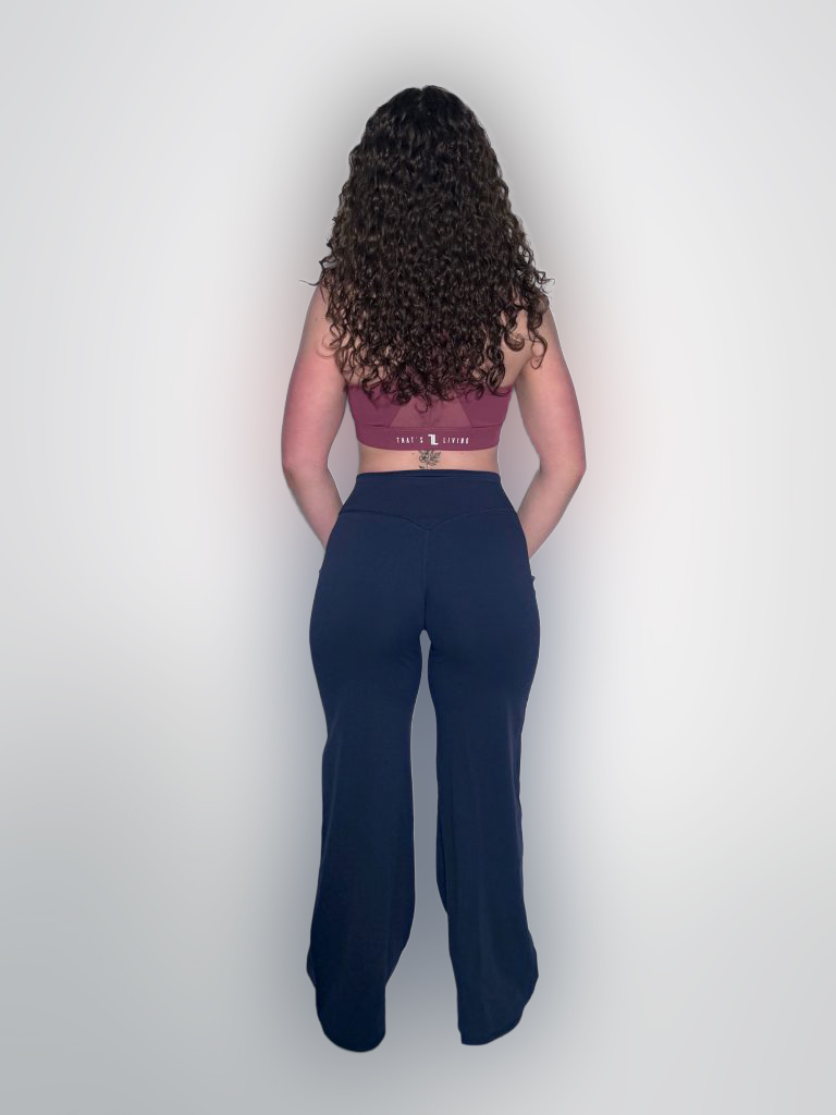 A person with long curly hair stands facing away, wearing a maroon crop top and black high-waisted pants, against a plain white background.