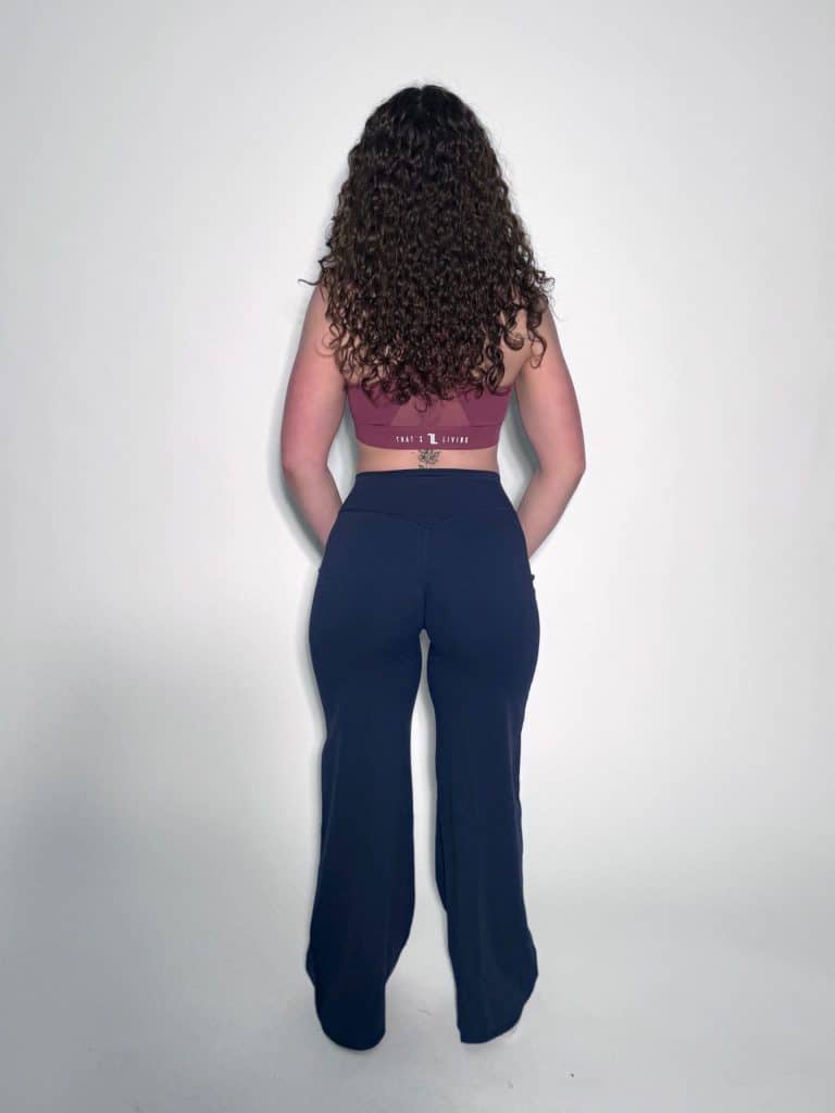 A person with long curly hair stands facing away, wearing a maroon crop top and black high-waisted pants, against a plain white background.