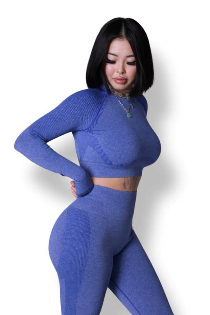 A woman with short black hair wears a matching blue long-sleeve crop top and high-waisted leggings. She stands against a white background, looking down with one hand resting on her back.