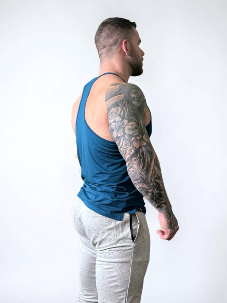 A muscular man wearing a blue tank top and gray sweatpants stands sideways, showing a full sleeve tattoo on his left arm. The background is plain and light-colored.