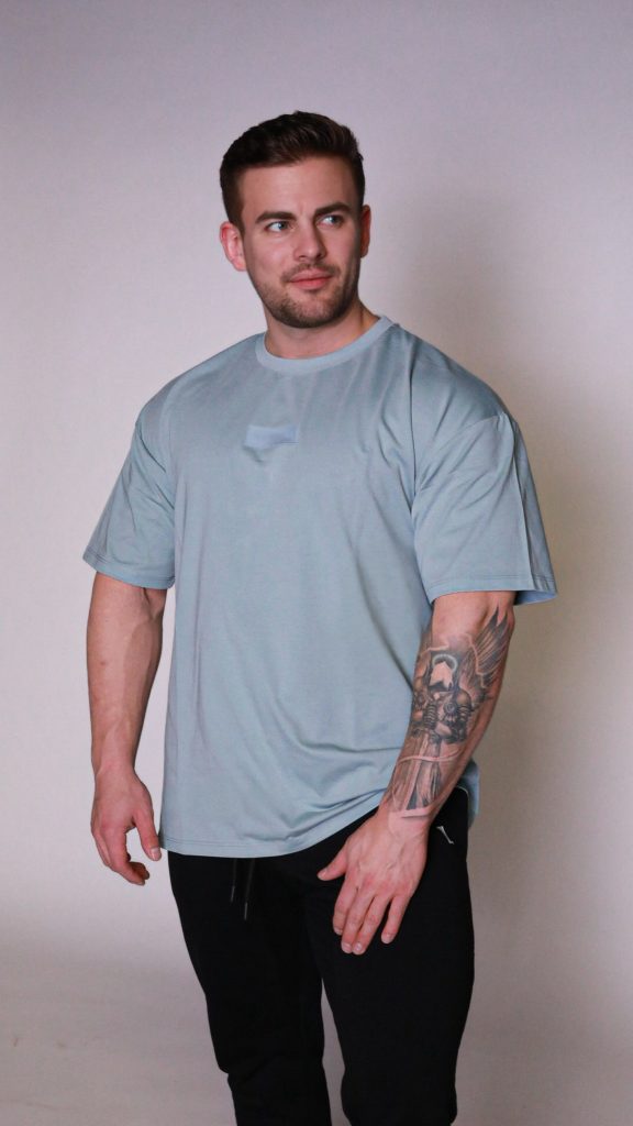 A man with short brown hair, a beard, and a tattoo sleeve on his left arm stands against a plain background, wearing the light blue Equity Tee with a tapered fit and scoop bottom, paired with black pants. He looks slightly to his right.