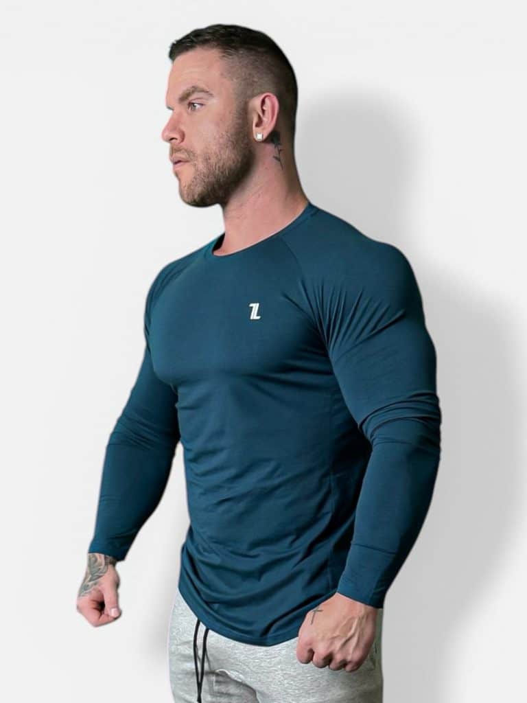 A man with short hair and a beard wears a fitted, long-sleeve teal athletic shirt with a small white "Z" logo on the chest and light gray pants, standing against a plain white background.