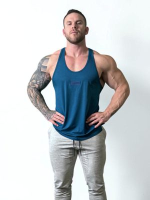 A muscular man with tattoos on his right arm stands confidently against a plain white background, wearing a blue tank top and light gray jogger pants, with his hands resting on his hips.
