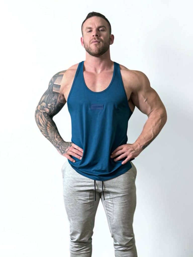 A muscular man with tattoos on his right arm stands confidently against a plain white background, wearing a blue tank top and light gray jogger pants, with his hands resting on his hips.