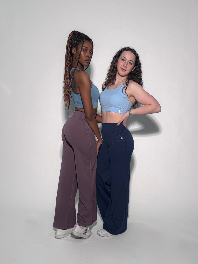 Two women stand confidently side by side in Sculpted Straight Flare Legging activewear—one in mauve, one in navy—paired with matching blue crop tops and white sneakers against a light background.