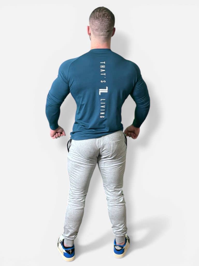 A muscular man stands facing away from the camera, wearing a fitted teal long-sleeve shirt with "That’s Living" text on the back, light gray pants, and blue and white sneakers against a plain white background.