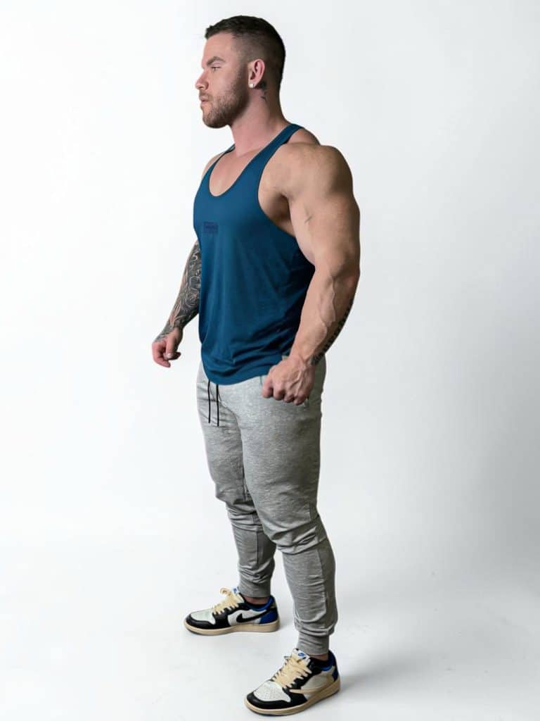 A muscular man with tattoos on his left arm stands in profile, wearing a blue tank top, light gray joggers, and sneakers, against a plain white background.