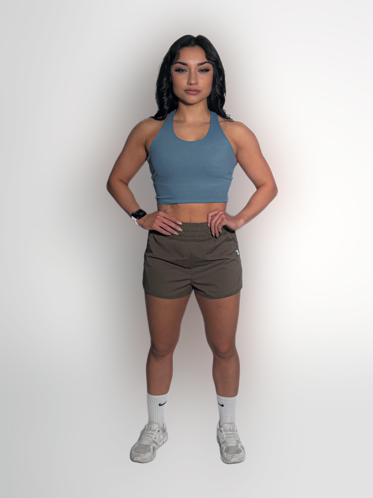 A woman stands against a plain white background, wearing a blue crop top, brown athletic shorts, white socks, and white sneakers. Her hands rest on her hips, and she has dark hair styled in loose waves.