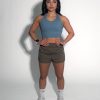 A woman stands against a plain white background, wearing a blue crop top, brown athletic shorts, white socks, and white sneakers. Her hands rest on her hips, and she has dark hair styled in loose waves.