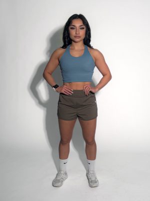 A woman stands against a plain white background, wearing a blue crop top, brown athletic shorts, white socks, and white sneakers. Her hands rest on her hips, and she has dark hair styled in loose waves.