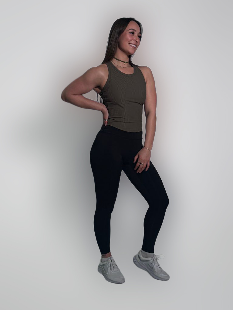 A smiling woman poses confidently in the LuxeForm Tank, paired with black leggings and sneakers, against a plain light background with her hand on her hip.