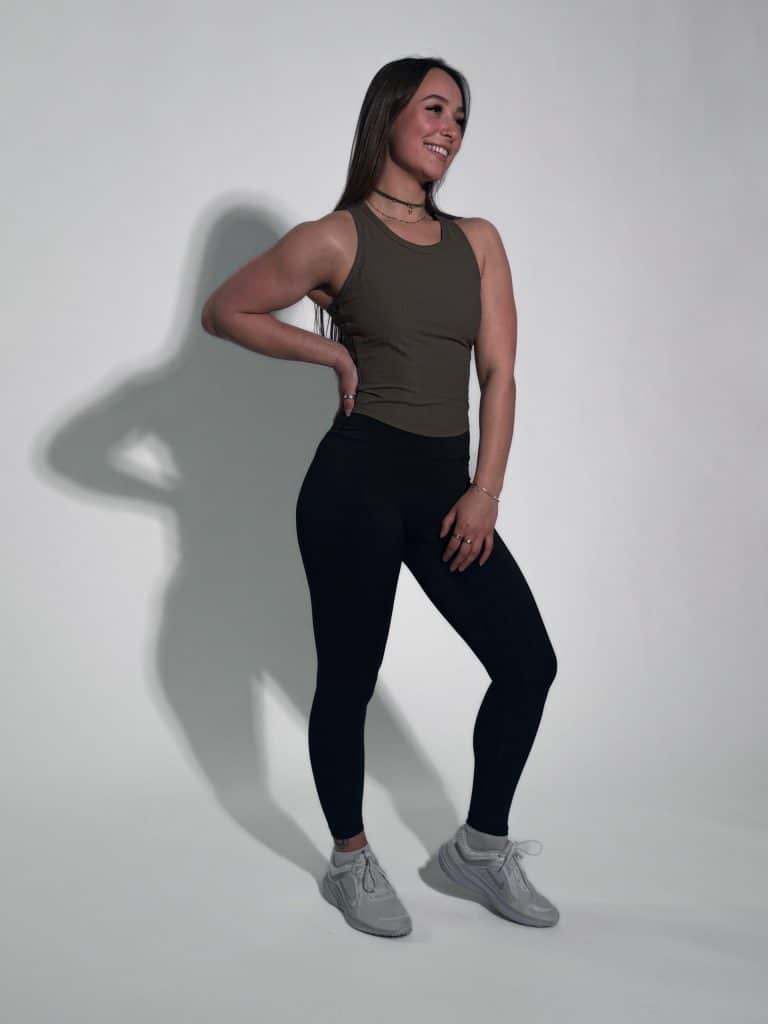 A smiling woman poses confidently in the LuxeForm Tank, paired with black leggings and sneakers, against a plain light background with her hand on her hip.