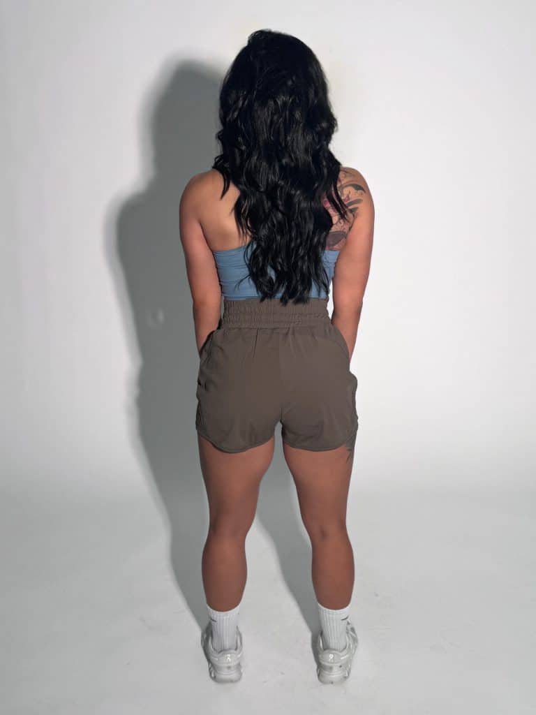 A woman with long dark wavy hair stands facing away from the camera. She is wearing a blue crop top, high-waisted olive shorts, white socks, and white sneakers against a plain light background.