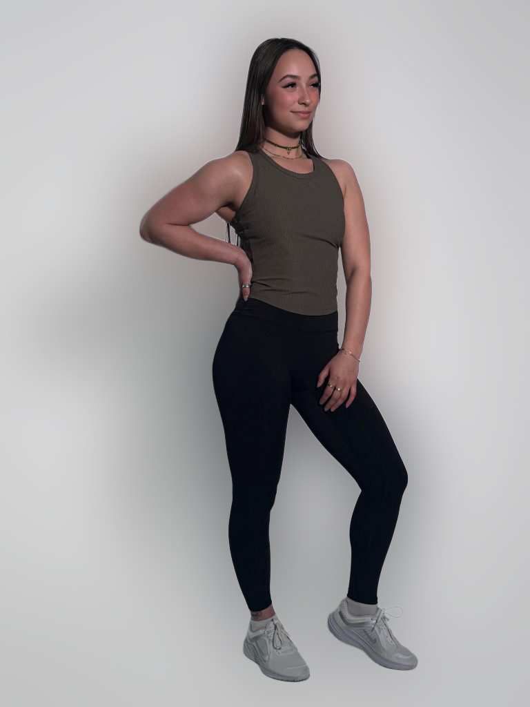 A woman wears the LuxeForm Tank in olive, paired with black leggings and gray sneakers, standing against a plain light background. She poses with one hand on her hip, looking slightly to the side and smiling.