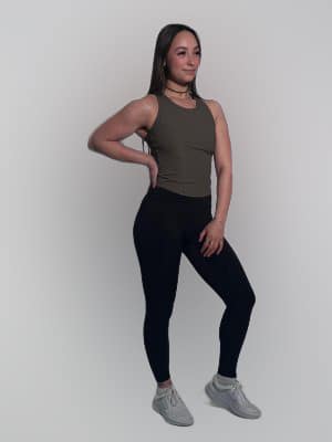 A woman wears the LuxeForm Tank in olive, paired with black leggings and gray sneakers, standing against a plain light background. She poses with one hand on her hip, looking slightly to the side and smiling.