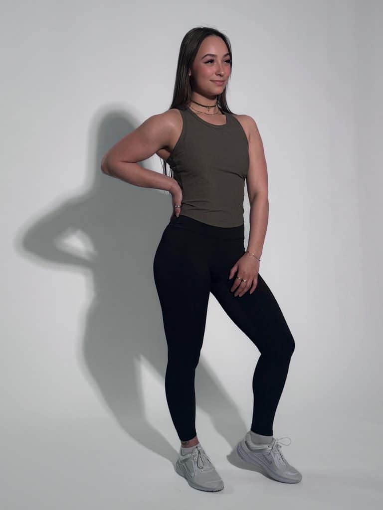 A woman wears the LuxeForm Tank in olive, paired with black leggings and gray sneakers, standing against a plain light background. She poses with one hand on her hip, looking slightly to the side and smiling.