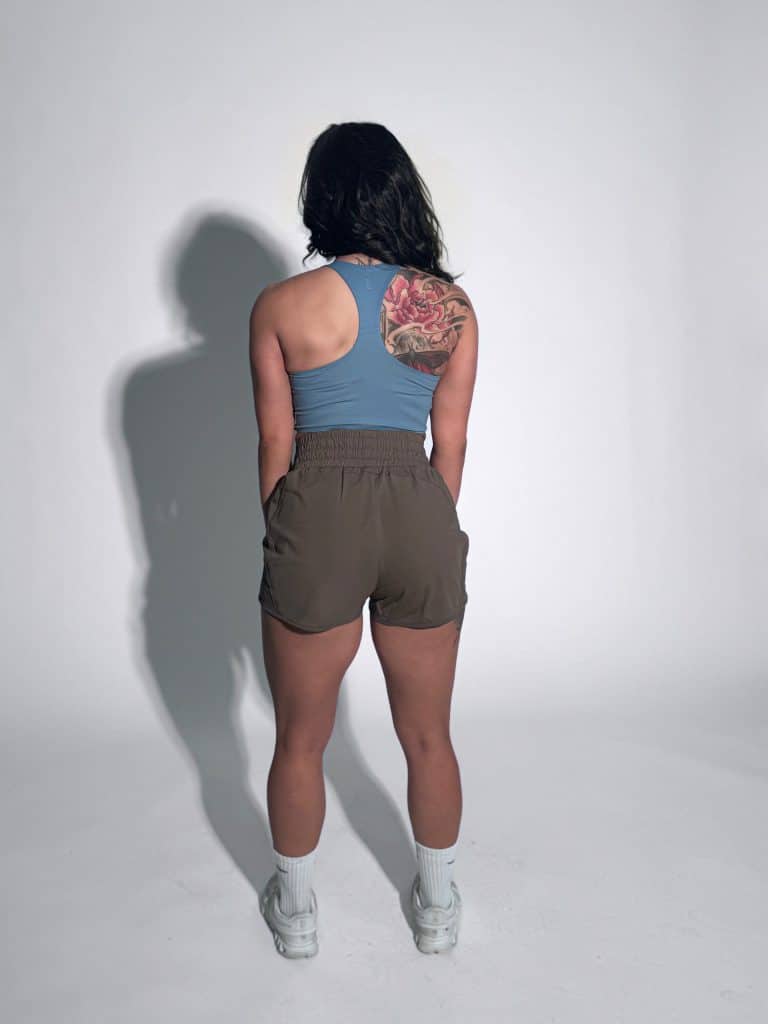 A woman stands facing away from the camera, wearing a blue tank top, brown shorts, white socks, and sneakers. She has a large, colorful floral tattoo on her right shoulder. The background is plain and white.