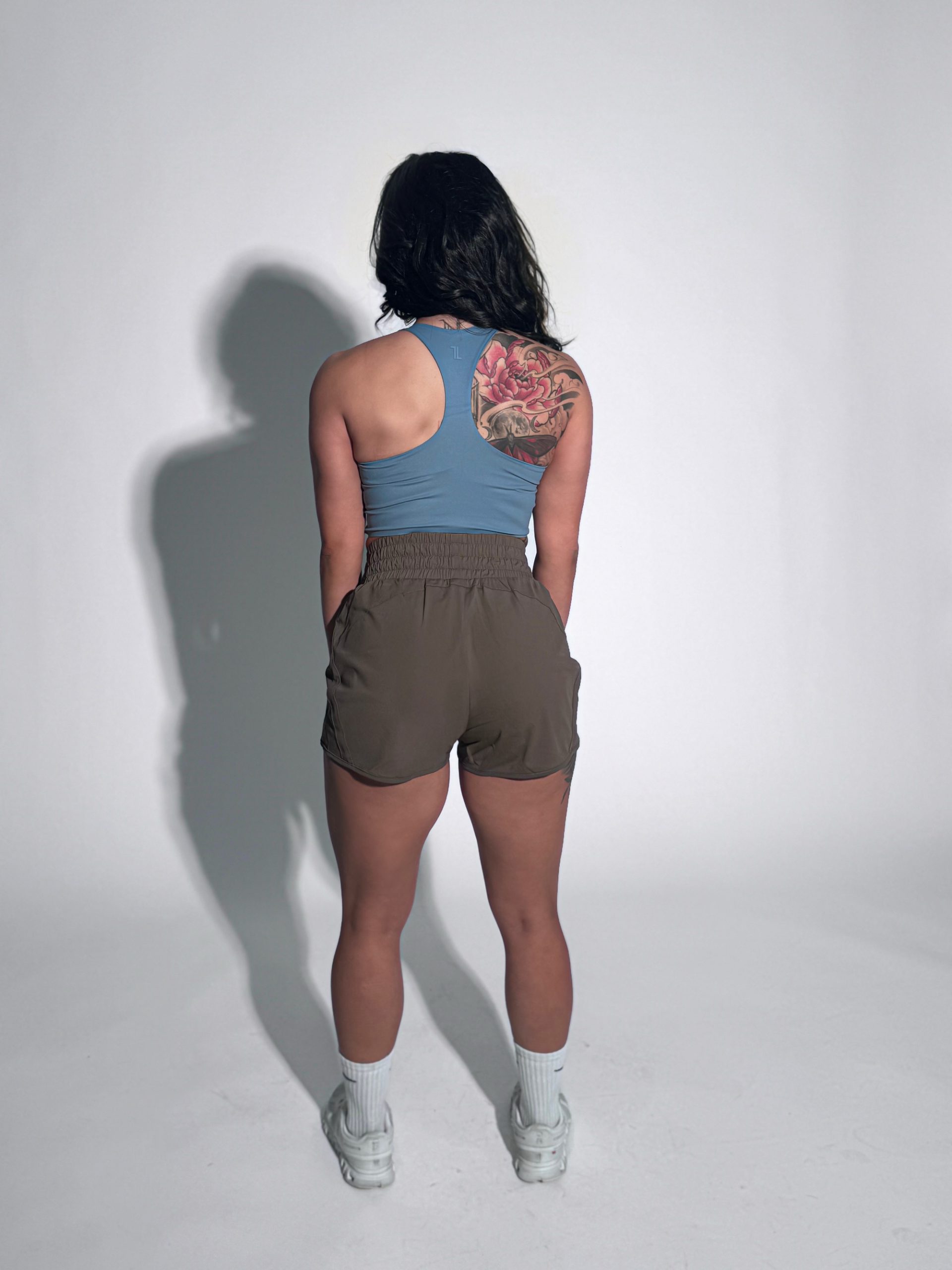 A woman stands facing away from the camera, wearing a blue tank top, brown shorts, white socks, and sneakers. She has a large, colorful floral tattoo on her right shoulder. The background is plain and white.