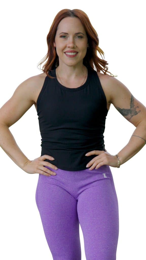 A woman with red hair smiles confidently, standing with her hands on her hips. She is wearing a sleeveless black top and purple leggings, with a visible tattoo on her left arm. The background is white.
