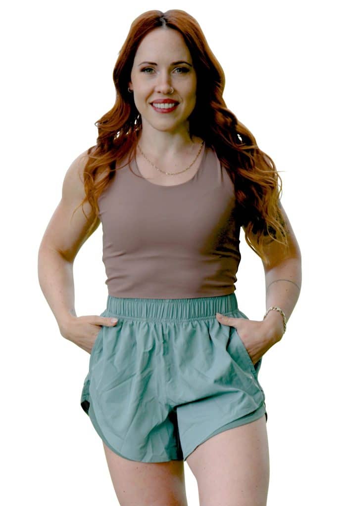 A woman with long red hair, wearing a sleeveless brown top and light green shorts, stands facing the camera with hands in her pockets and a friendly smile.