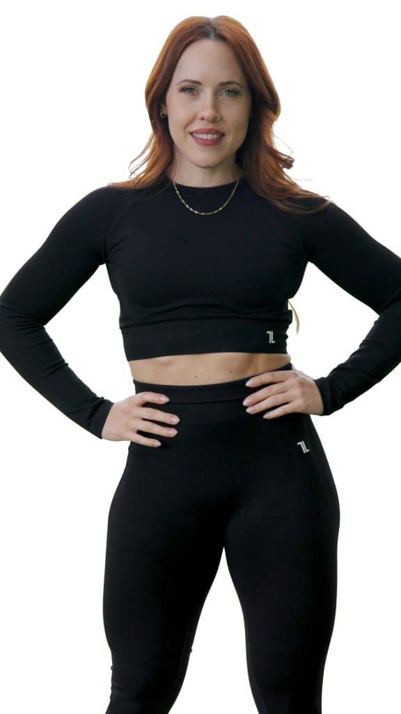 A woman with long red hair stands confidently with hands on hips, wearing a black long-sleeve crop top and matching high-waisted leggings, smiling against a plain white background.