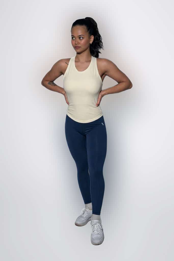 A woman stands against a plain background with hands on hips, wearing a cream tank top, blue leggings, and white sneakers. She has a tattoo on her right arm and her dark hair is tied back in a ponytail.