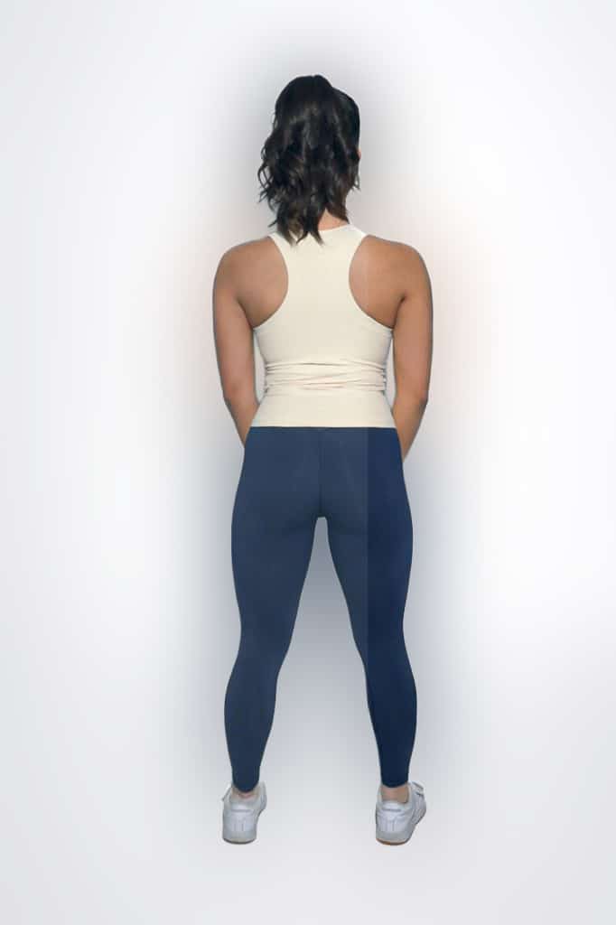 A woman stands facing away from the camera against a white background, wearing a sleeveless beige top, navy blue leggings, and white sneakers. Her arms are relaxed by her sides.