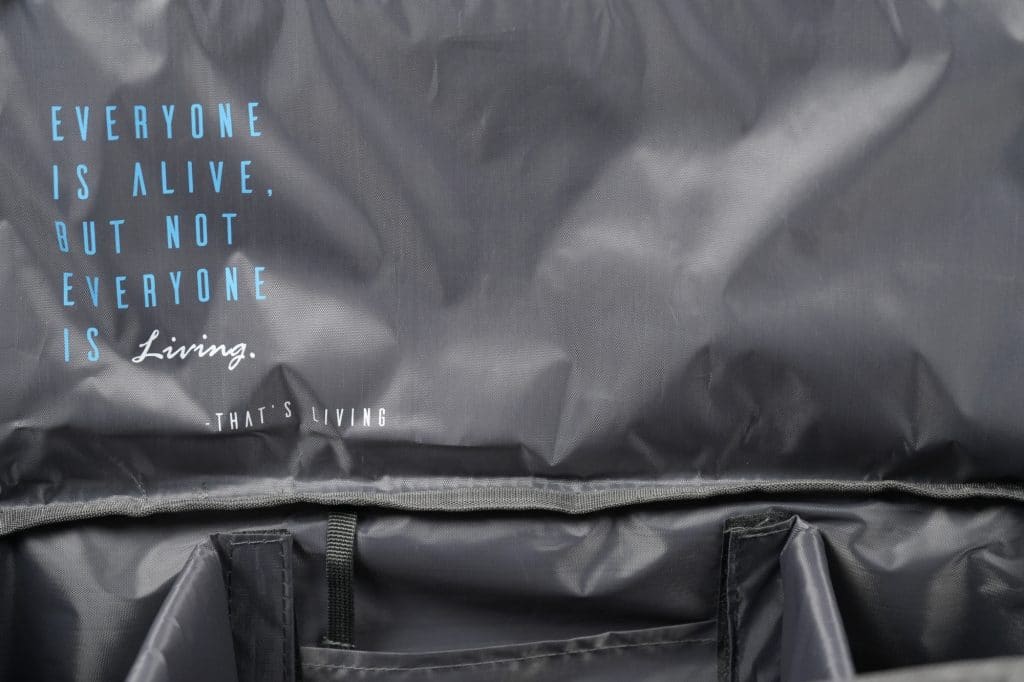 The TRIARC Duffle Bag is dark gray with the quote "Everyone is alive, but not everyone is living. That’s living" in blue and white text. The photo also shows its spacious inner compartments.
