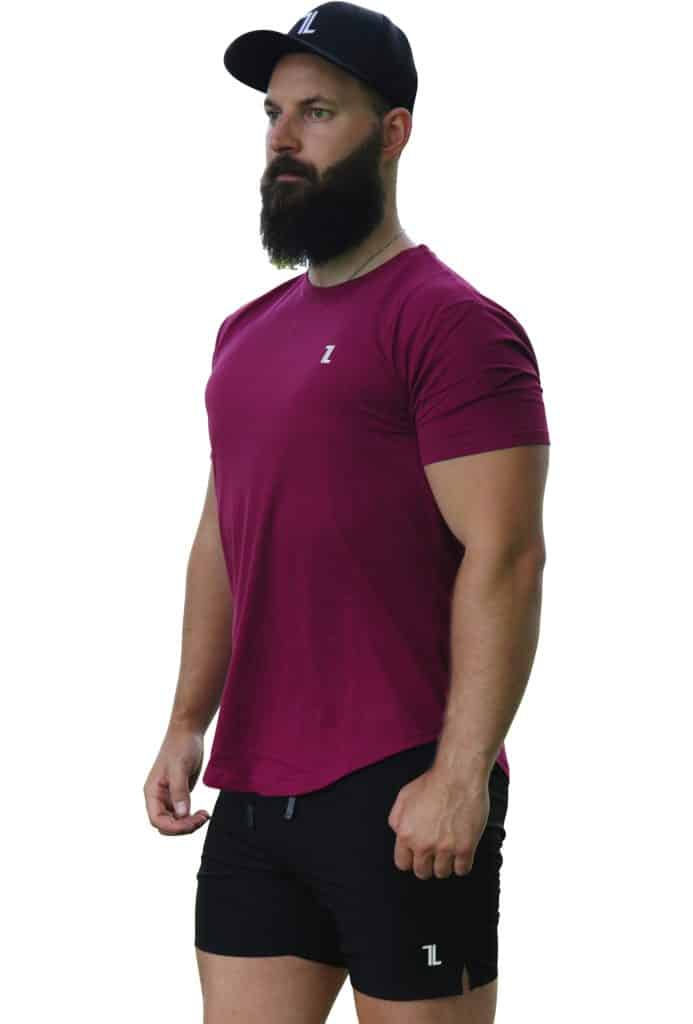 A man with a beard wears a black cap, a maroon T-shirt, and black shorts with a small white logo, standing against a white background.