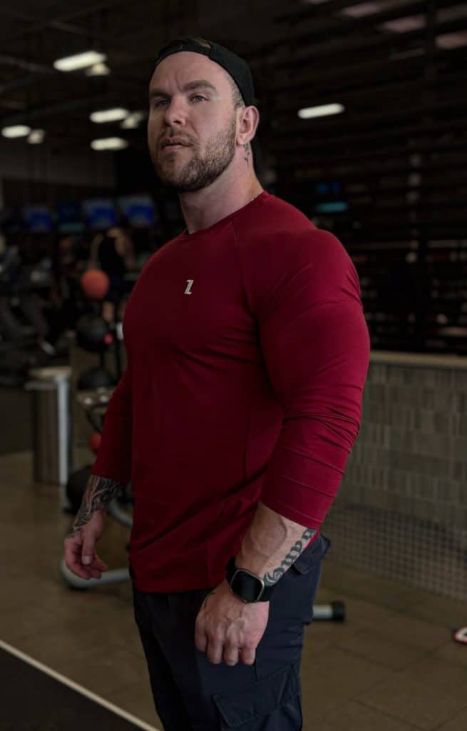 A muscular man with tattoos, wearing a red FRMR Long sleeve shirt, black pants, a black smartwatch, and a backward black cap stands in a gym with exercise equipment in the background.