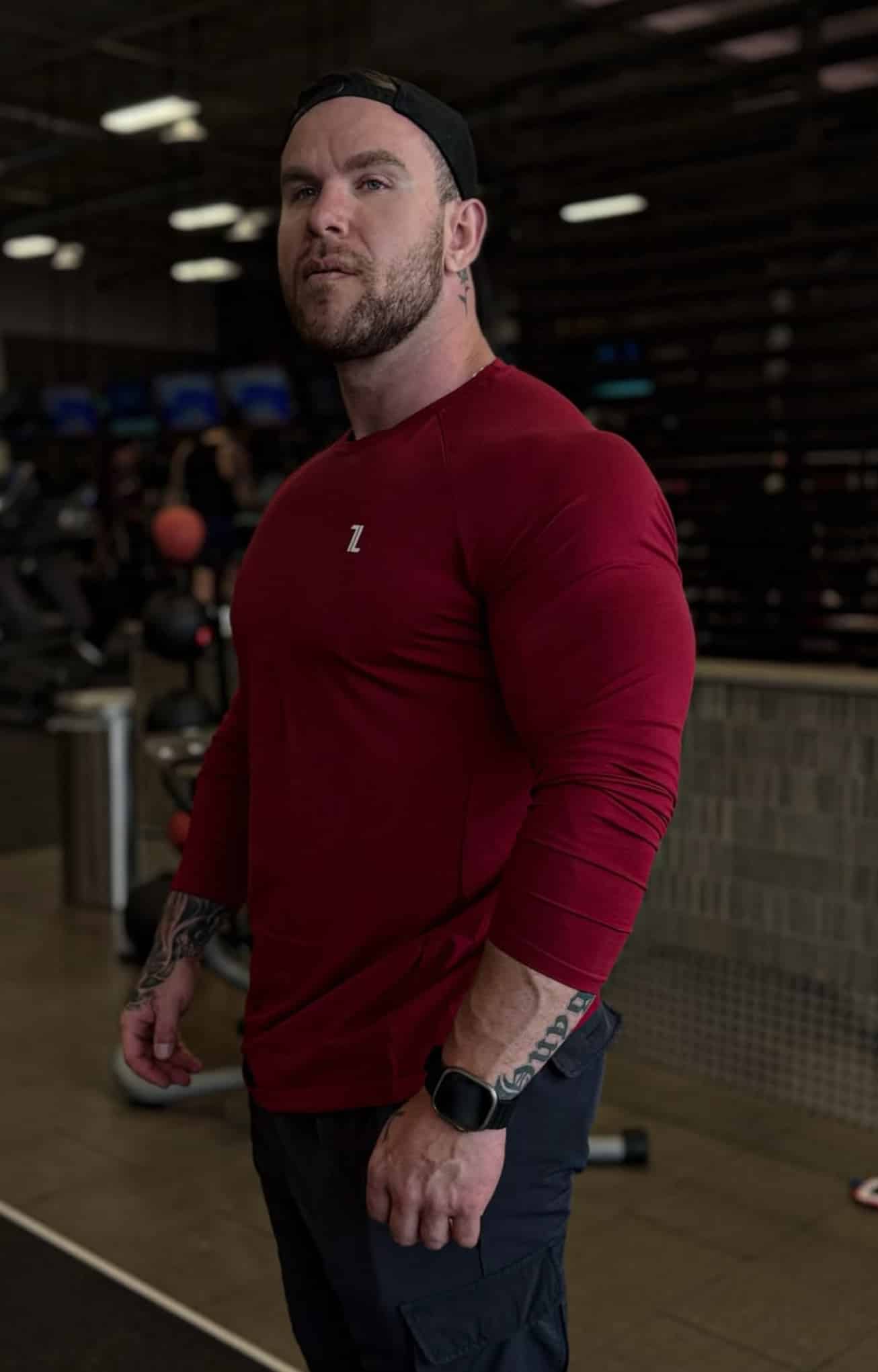 A muscular man with tattoos, wearing a red FRMR Long sleeve shirt, black pants, a black smartwatch, and a backward black cap stands in a gym with exercise equipment in the background.