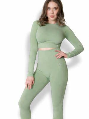 A woman models the Allure Set V2—a fitted, long-sleeve sage green crop top with matching high-waisted leggings—against a plain white background, posing with one hand on her hip and the other by her side.