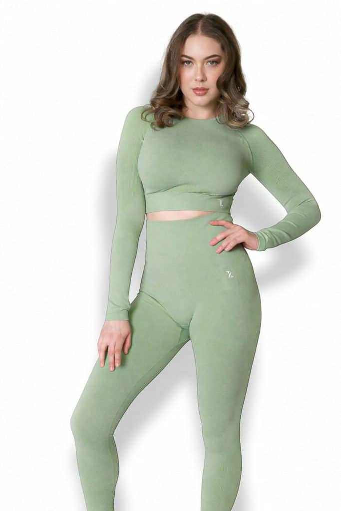 A woman models the Allure Set V2—a fitted, long-sleeve sage green crop top with matching high-waisted leggings—against a plain white background, posing with one hand on her hip and the other by her side.