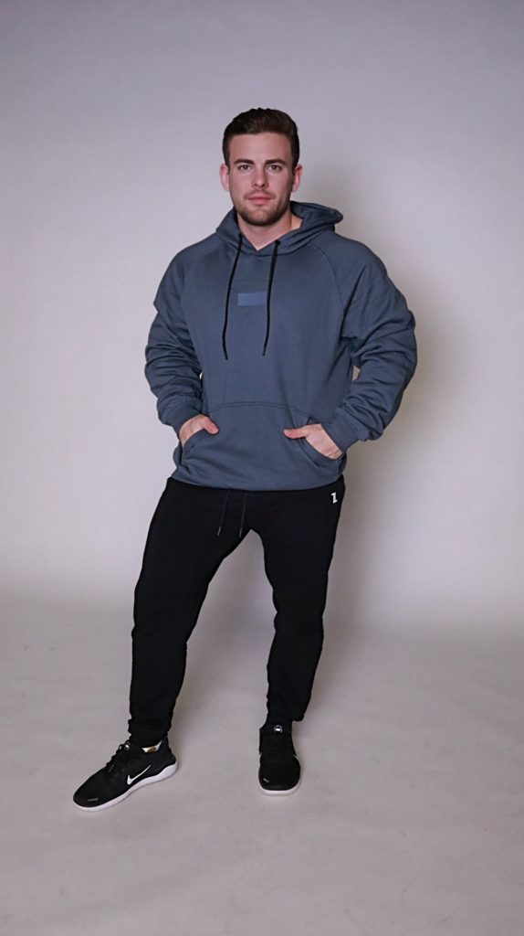 A man poses against a light background, hands in his pockets, wearing the blue FORGE HOODIE over a men's performance tee, paired with black jogger pants and sneakers. He stands facing forward in a casual, stylish look.