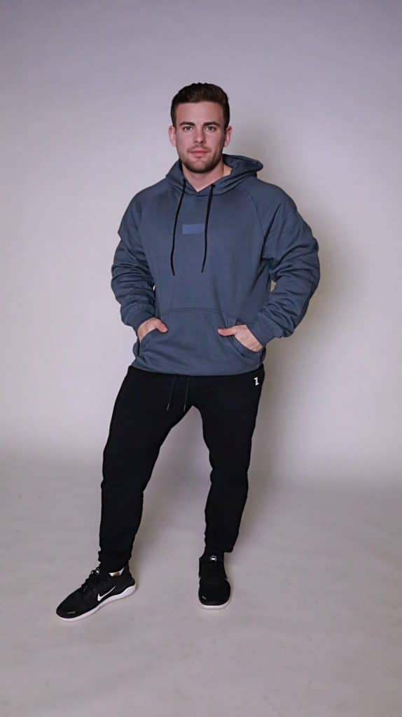 A man poses against a light background, hands in his pockets, wearing the blue FORGE HOODIE over a men's performance tee, paired with black jogger pants and sneakers. He stands facing forward in a casual, stylish look.