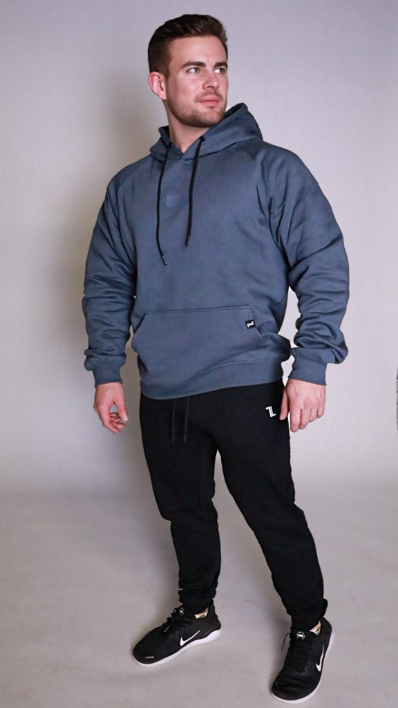 A man stands in a studio wearing a blue-gray hoodie, black jogger pants, and black sneakers. He looks slightly to the side with a neutral expression, and the background is plain light gray.