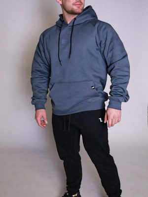 A man stands in a studio wearing a blue-gray hoodie, black jogger pants, and black sneakers. He looks slightly to the side with a neutral expression, and the background is plain light gray.