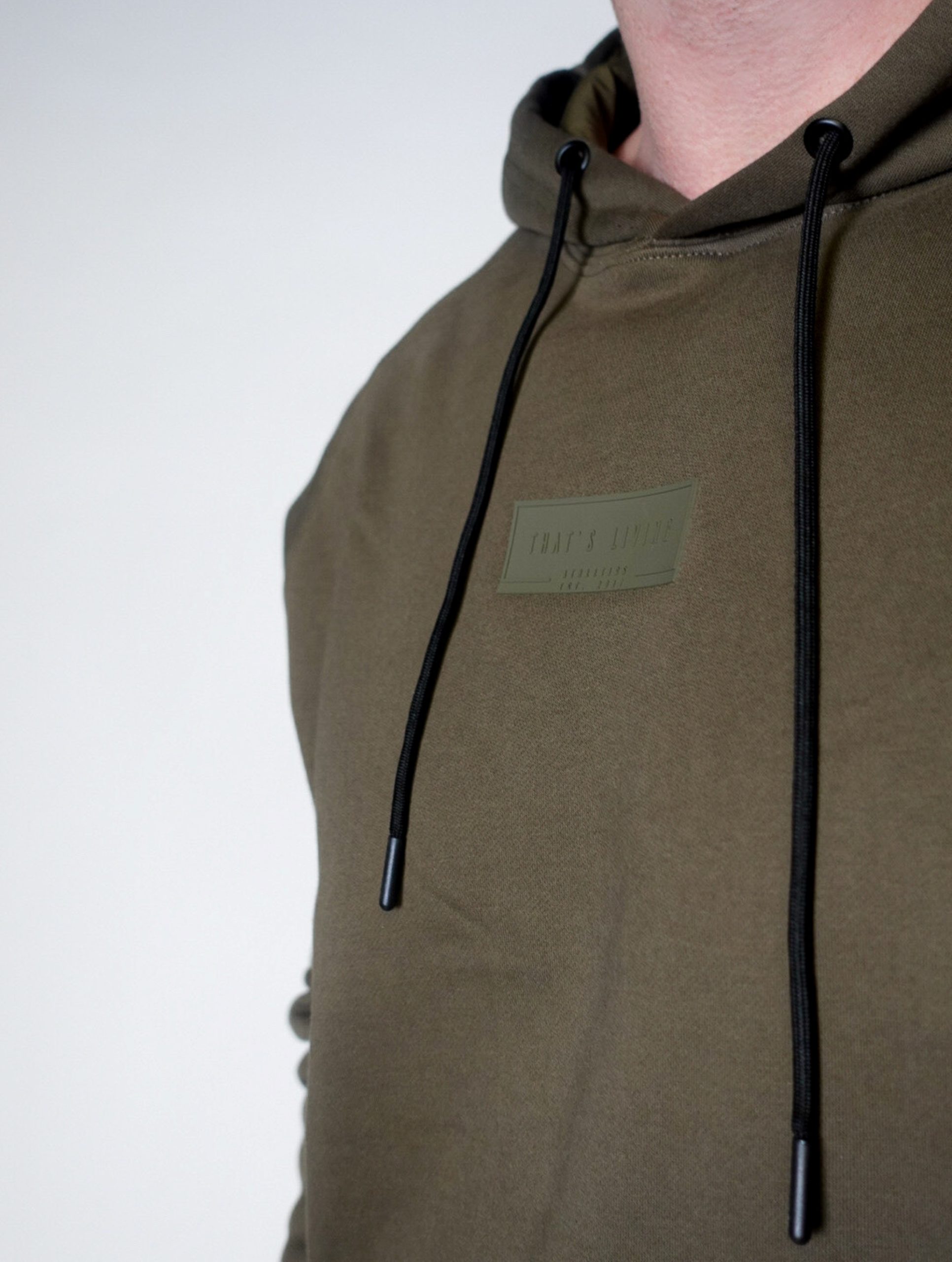 A person wearing an olive green hoodie with black drawstrings. The hoodie has a rectangular patch on the chest with some text. The person's face is partially out of the frame.