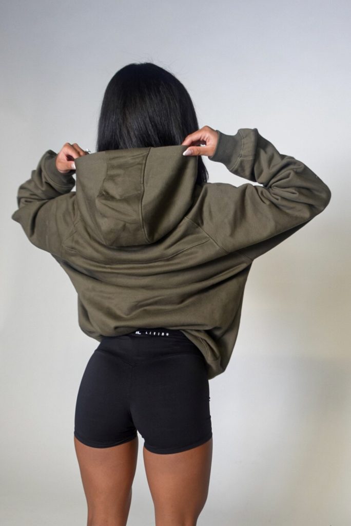 A person with long dark hair, seen from behind, adjusts the hood of an olive green hoodie while wearing black fitted shorts against a plain, light background.