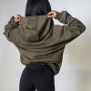 Forge Olive 2 A person with long dark hair, seen from behind, adjusts the hood of an olive green hoodie while wearing black fitted shorts against a plain, light background.