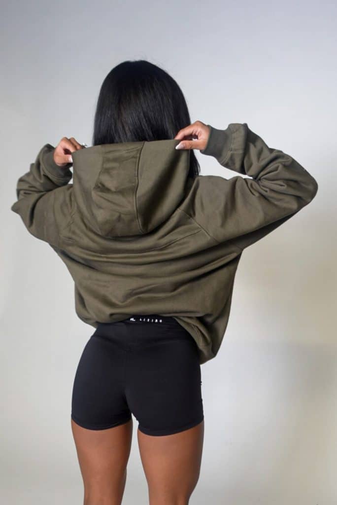 A person with long dark hair, seen from behind, adjusts the hood of an olive green hoodie while wearing black fitted shorts against a plain, light background.