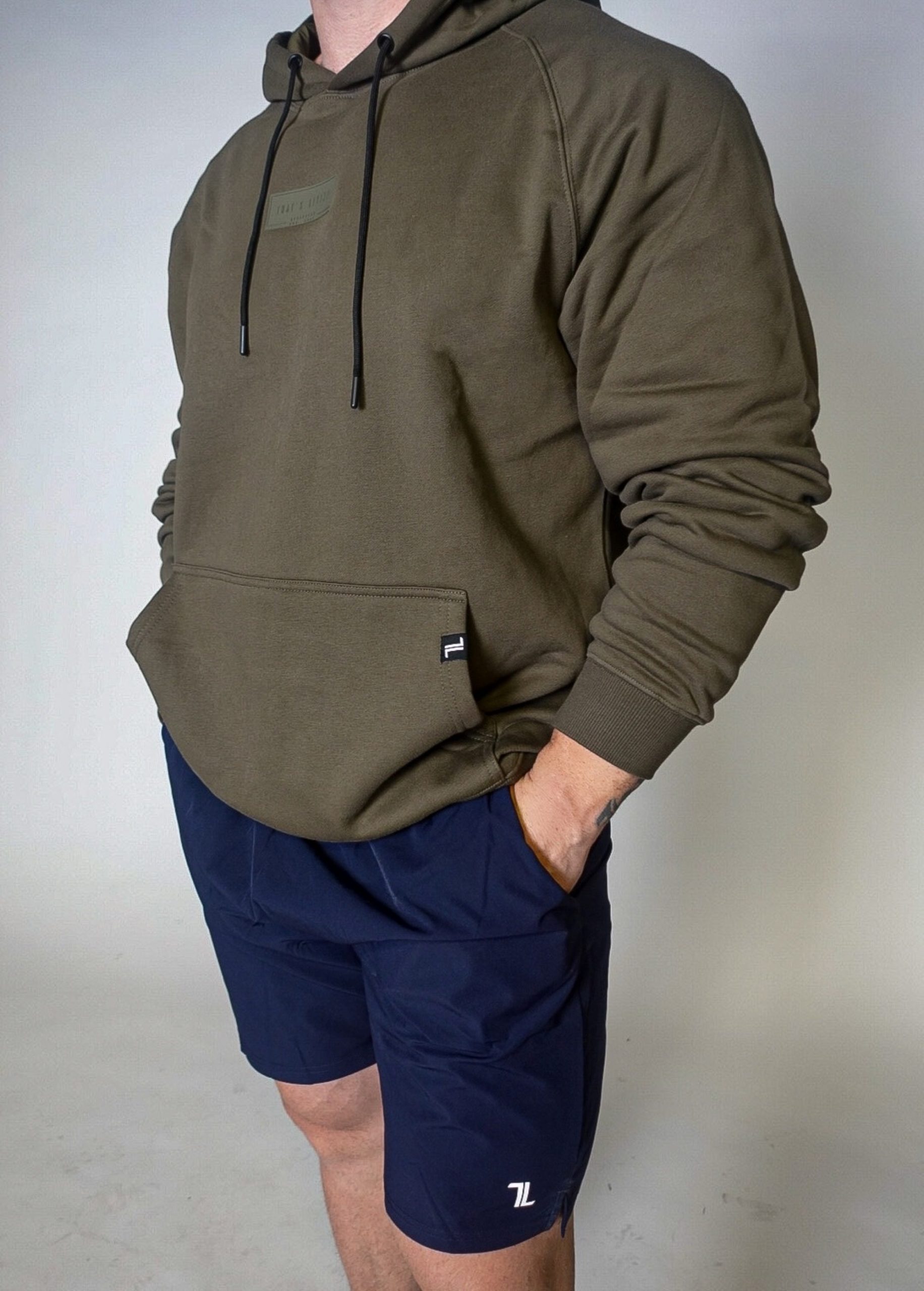 A person wearing an olive green hoodie with a front pocket and navy blue shorts, standing with one hand in their pocket against a plain, light background. Their face is not visible.