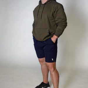 A man stands against a plain light background, wearing an olive green hoodie, navy blue shorts, and black sneakers. His hands are in his pockets, and he looks slightly upward with a neutral expression.